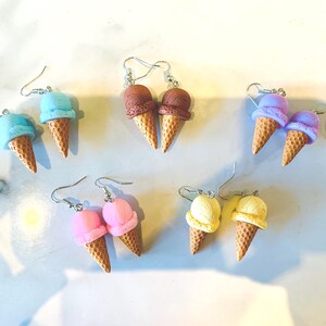 K&ouml;nnte beinhalten: Eine Sammlung farbenfroher Eiscreme-Kegel-Ohrringe. Die Ohrringe zeigen Kugeln aus blauer, brauner, lila, rosa und gelber Eiscreme auf strukturierten Kegelbasen. Die Ohrringe werden auf einer wei&szlig;en Oberfl&auml;che pr&auml;sentiert.