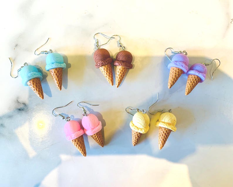 K&ouml;nnte beinhalten: Eine Sammlung farbenfroher Eiscreme-Kegel-Ohrringe. Die Ohrringe zeigen Kugeln aus blauer, brauner, lila, rosa und gelber Eiscreme auf strukturierten Kegelbasen. Die Ohrringe werden auf einer wei&szlig;en Oberfl&auml;che pr&auml;sentiert.