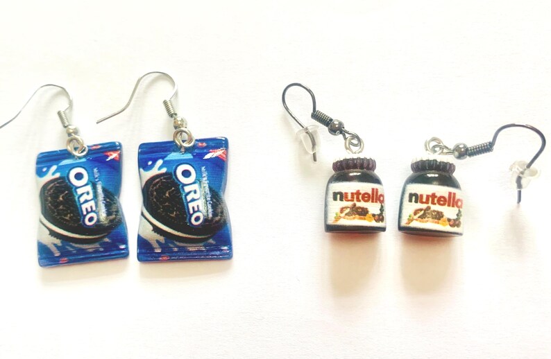 K&ouml;nnte beinhalten: Ohrringe mit Miniatur-Lebensmitteln. Ein Paar zeigt Oreo-Keksverpackungen in Blau und Wei&szlig;, mit einem schwarzen Keks. Das andere Paar zeigt kleine Nutella-Gl&auml;ser mit braunen Deckeln und wei&szlig;en Etiketten.