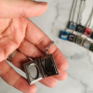 May include: Silver-toned locket necklace, open to reveal two oval spaces for photos. The locket hangs from a delicate chain. Other similar necklaces are visible in the background, each with a small, square pendant.