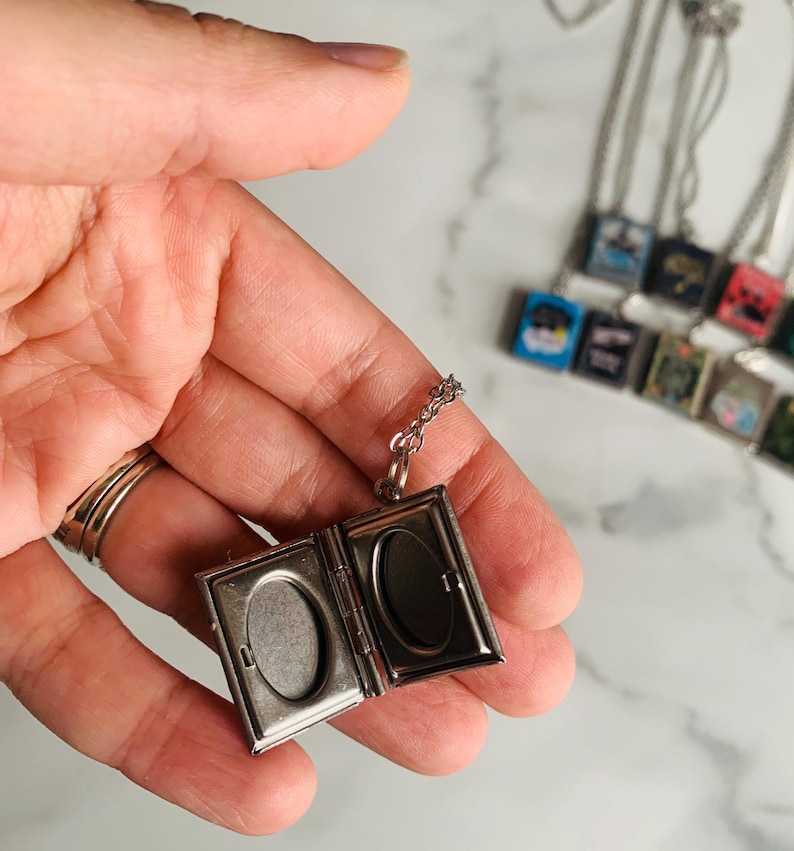 May include: Silver-toned locket necklace, open to reveal two oval spaces for photos. The locket hangs from a delicate chain. Other similar necklaces are visible in the background, each with a small, square pendant.