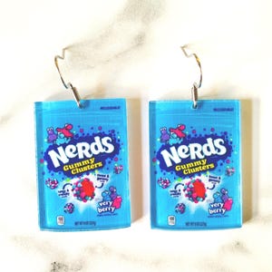 K&ouml;nnte beinhalten: Zwei blaue Acryl-Ohrringe, die wie Nerds Gummy Clusters-Bonbonpackungen geformt sind. Jeder Ohrring zeigt das Nerds-Logo, S&uuml;&szlig;igkeiten-Grafiken und den Schriftzug "very berry". Die Ohrringe haben silberfarbene Fischhaken-Ohrdr&auml;hte.