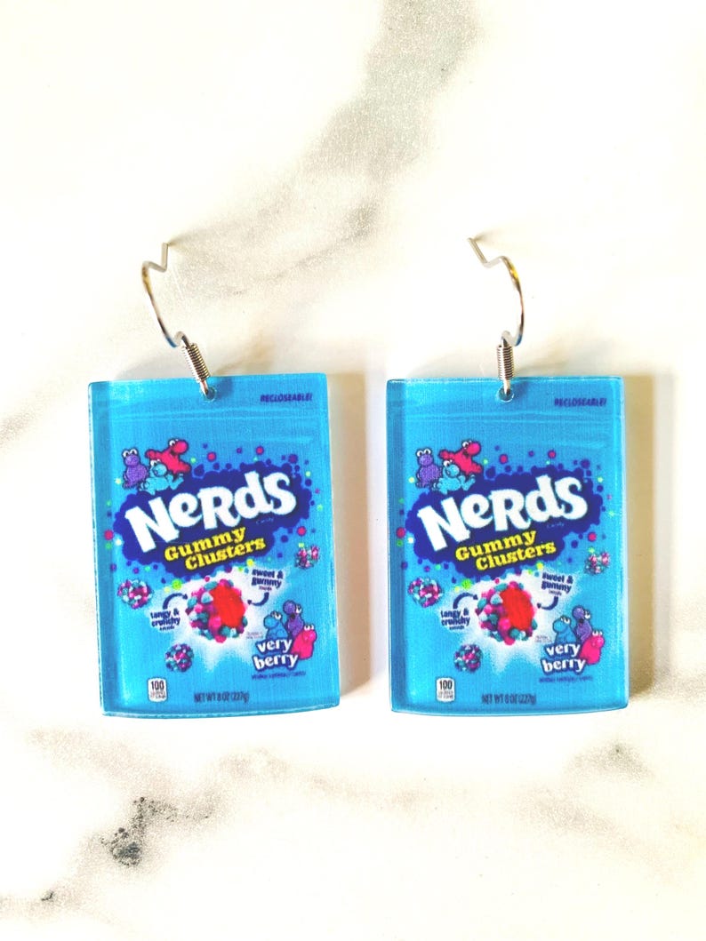 K&ouml;nnte beinhalten: Zwei blaue Acryl-Ohrringe, die wie Nerds Gummy Clusters-Bonbonpackungen geformt sind. Jeder Ohrring zeigt das Nerds-Logo, S&uuml;&szlig;igkeiten-Grafiken und den Schriftzug "very berry". Die Ohrringe haben silberfarbene Fischhaken-Ohrdr&auml;hte.
