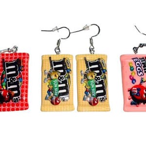 May include: A set of three pairs of dangle earrings featuring candy bar designs. The first pair is red with white polka dots and the text "M&M's". The second pair is yellow with the text "M&M's Fun Size". The third pair is pink with the text "M&M's Eggs".