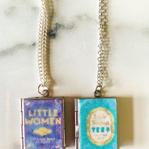 May include: Two silver chain necklaces each feature a small, book-shaped pendant. One pendant has a blue cover with the words "Little Women" in gold. The other pendant is turquoise with the same title.
