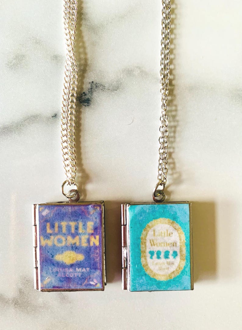 May include: Two silver chain necklaces each feature a small, book-shaped pendant. One pendant has a blue cover with the words "Little Women" in gold. The other pendant is turquoise with the same title.