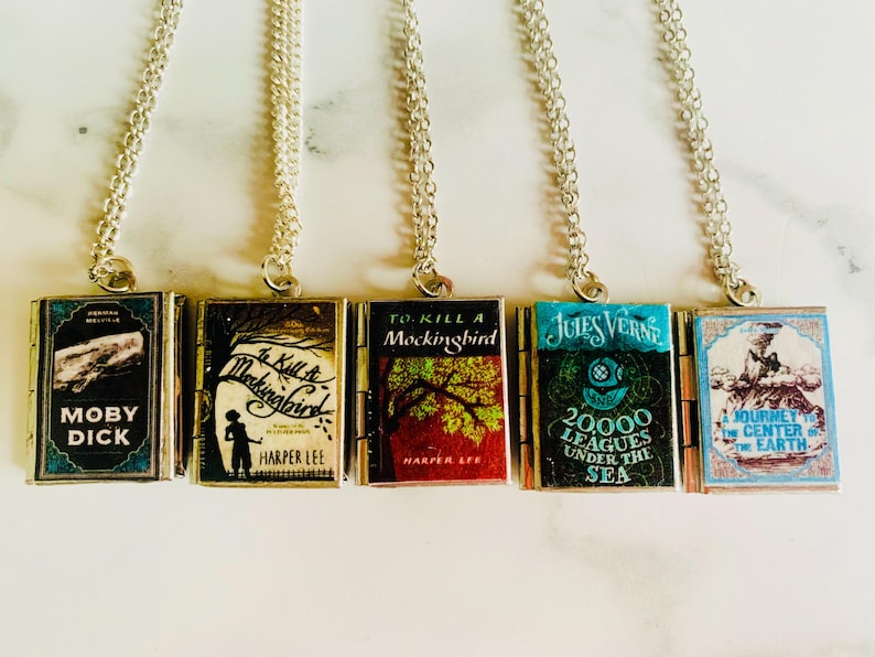 May include: Five silver-toned necklaces, each featuring a miniature book pendant. The book covers display titles like "Moby Dick" and "To Kill a Mockingbird." The pendants are rectangular with detailed cover art and text.