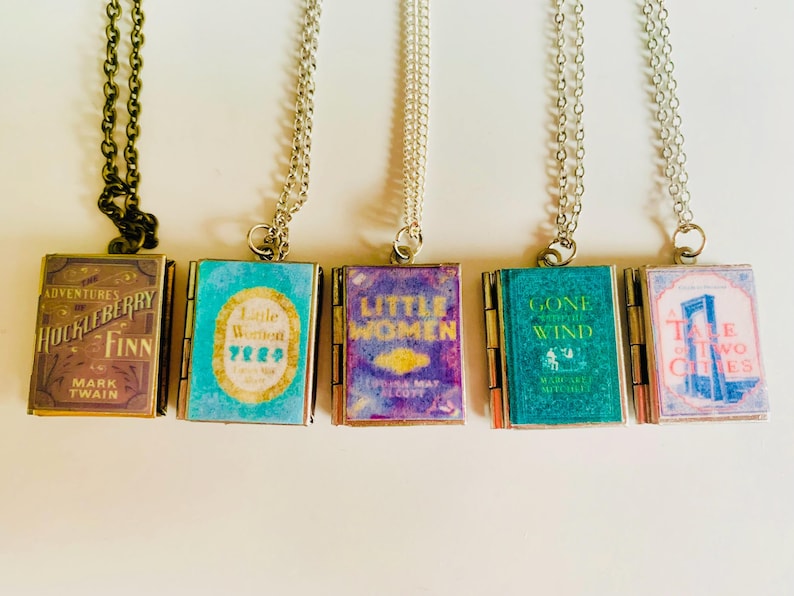 May include: Five book-shaped pendant necklaces. The pendants feature book covers from classic novels, including "Adventures of Huckleberry Finn" and "Little Women." Each pendant is attached to a chain. The first chain is dark bronze, and the others are silver.