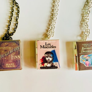 May include: Three pendant necklaces featuring miniature book covers. The first is a dark bronze chain with a pendant of "The Adventures of Huckleberry Finn." The second is a silver chain with "Les Mis&eacute;rables." The third is a silver chain with "Handbook for the Recently Deceased."