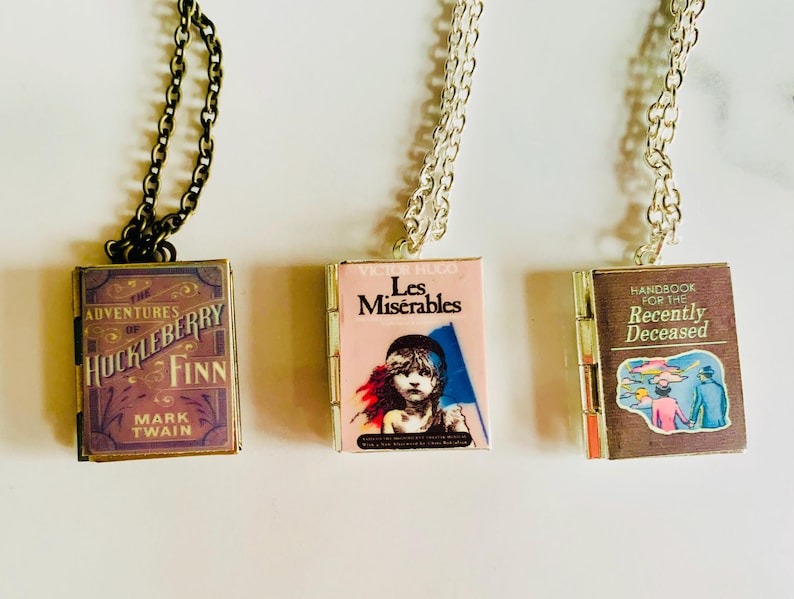 May include: Three pendant necklaces featuring miniature book covers. The first is a dark bronze chain with a pendant of "The Adventures of Huckleberry Finn." The second is a silver chain with "Les Mis&eacute;rables." The third is a silver chain with "Handbook for the Recently Deceased."