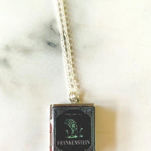 May include: Silver-toned necklace with a small, rectangular pendant featuring the cover art of "Frankenstein" by Mary Shelley. The pendant has a black background with a green illustration of a hand and the title in white.