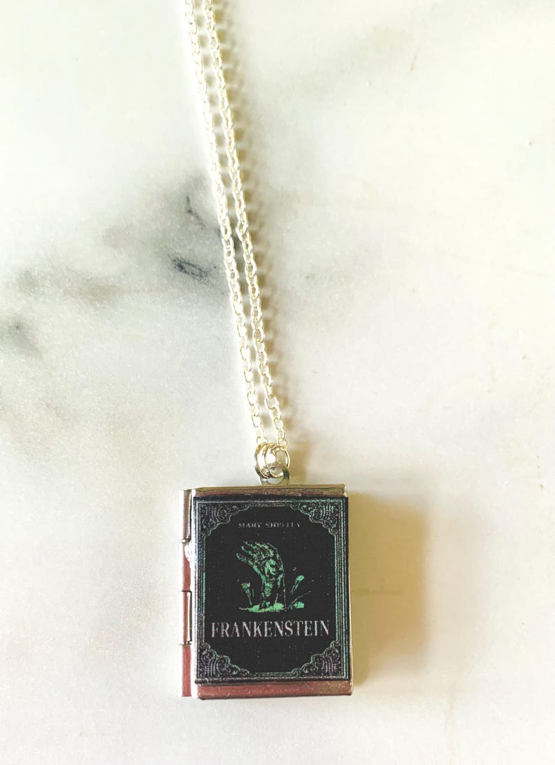 May include: Silver-toned necklace with a small, rectangular pendant featuring the cover art of "Frankenstein" by Mary Shelley. The pendant has a black background with a green illustration of a hand and the title in white.
