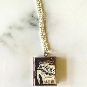 May include: A silver necklace with a rectangular pendant featuring the cover of "To Kill a Mockingbird" by Harper Lee. The pendant shows a silhouette of a child and a tree, with the book title in cursive.