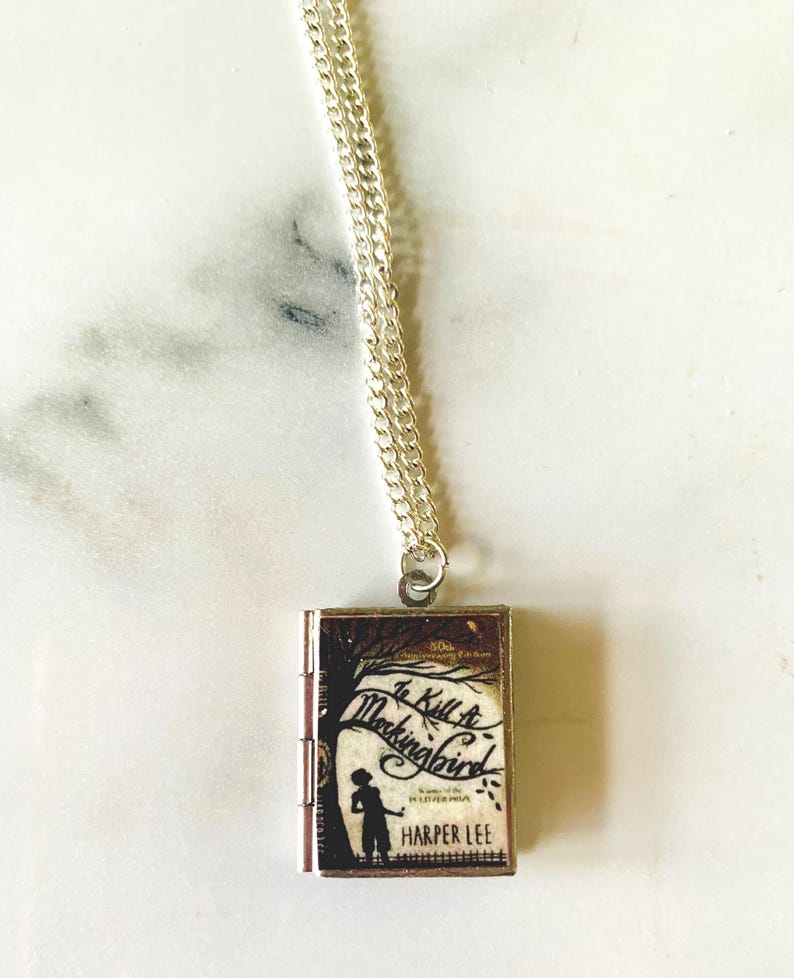May include: A silver necklace with a rectangular pendant featuring the cover of "To Kill a Mockingbird" by Harper Lee. The pendant shows a silhouette of a child and a tree, with the book title in cursive.