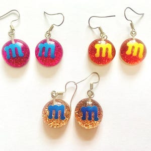 K&ouml;nnte beinhalten: F&uuml;nf Paar Ohrh&auml;nger mit glitzerndem, rundem Design. Die Ohrringe zeigen den Buchstaben "M" in Blau oder Gelb, vor einem rosa, roten oder goldenen Hintergrund. Die Ohrringe haben silberfarbene Haken.