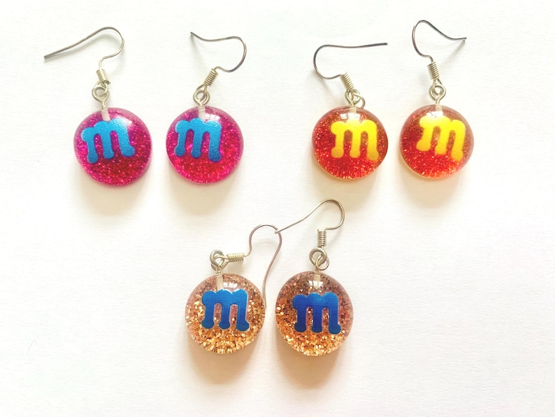 K&ouml;nnte beinhalten: F&uuml;nf Paar Ohrh&auml;nger mit glitzerndem, rundem Design. Die Ohrringe zeigen den Buchstaben "M" in Blau oder Gelb, vor einem rosa, roten oder goldenen Hintergrund. Die Ohrringe haben silberfarbene Haken.