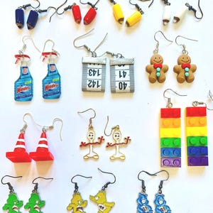 Może przedstawiać: Kolekcja kolczyków w różnych wzorach, w tym ołówki, czerwone pachołki drogowe, miarki, piernikowe ludziki, tęczowe klocki i Misie Tulisie. Kolczyki są prezentowane na białej powierzchni, prezentując różnorodność kolorów i stylów.