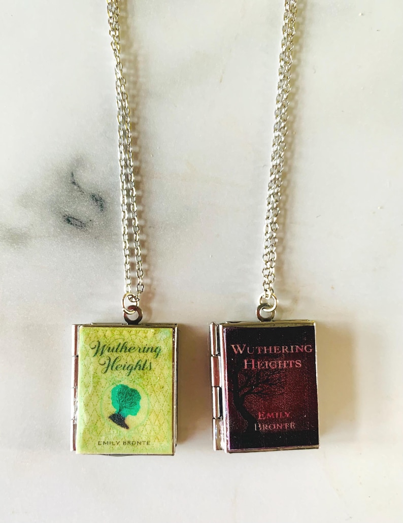 May include: Two silver chain necklaces each feature a small, book-shaped pendant. One pendant is green and yellow with the title "Wuthering Heights" and a silhouette. The other is burgundy with the same title.