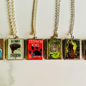 May include: Six silver-toned necklaces, each featuring a miniature book pendant. The book covers display titles like "Handbook for the Recently Deceased", "Inferno", and "Pet Sematary". The pendants are connected to delicate chains.