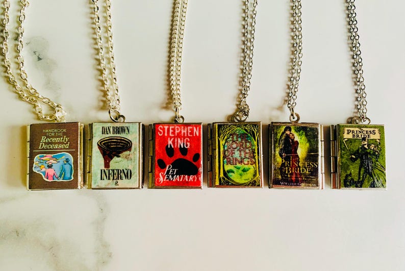 May include: Six silver-toned necklaces, each featuring a miniature book pendant. The book covers display titles like "Handbook for the Recently Deceased", "Inferno", and "Pet Sematary". The pendants are connected to delicate chains.