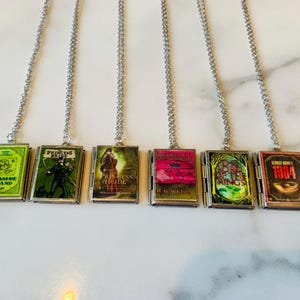May include: Six silver-toned necklaces each feature a miniature book pendant. The book covers display titles like "Treasure Island" and "1984." The pendants are rectangular with a hinged design, and the book covers are colorful.