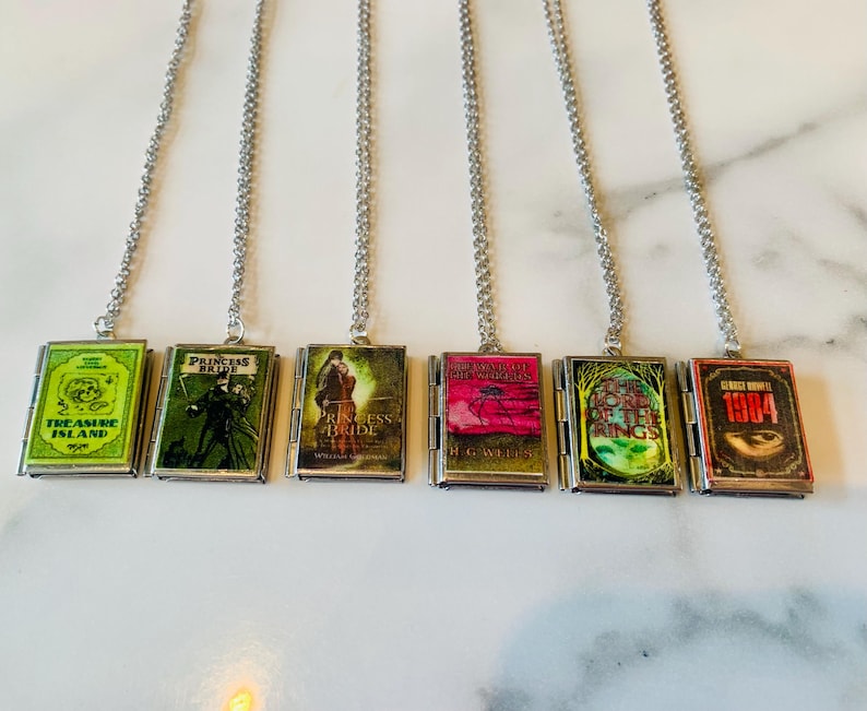 May include: Six silver-toned necklaces each feature a miniature book pendant. The book covers display titles like "Treasure Island" and "1984." The pendants are rectangular with a hinged design, and the book covers are colorful.