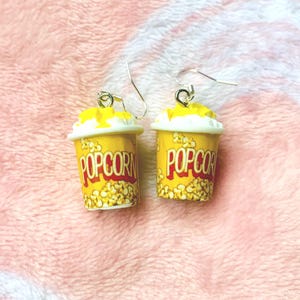 K&ouml;nnte beinhalten: Ein Paar ausgefallene Ohrringe in Form von Popcorn-Beh&auml;ltern. Die Ohrringe zeigen gelbe Beh&auml;lter mit dem Wort "POP CORN" in Rot, gef&uuml;llt mit Popcorn und mit gelber Butter belegt. Die Ohrringe h&auml;ngen an silberfarbenen Haken.