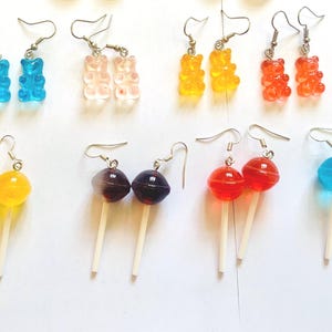 K&ouml;nnte beinhalten: Eine Sammlung bunter Modeschmuck-Ohrringe. Die Ohrringe zeigen verschiedene Designs, darunter Gummib&auml;rchen, Lutscher und runde S&uuml;&szlig;igkeiten in Blau, Rosa, Gelb, Orange, Rot und Lila. Jedes Paar hat einen silbernen Haken.