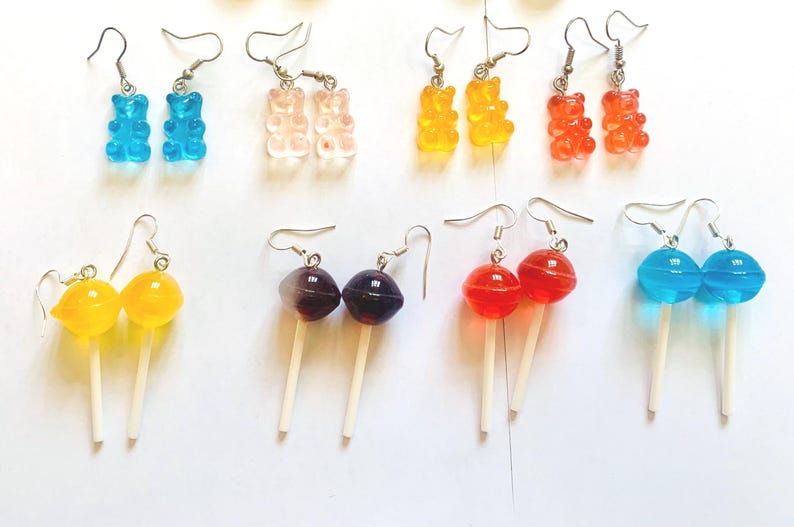 K&ouml;nnte beinhalten: Eine Sammlung bunter Modeschmuck-Ohrringe. Die Ohrringe zeigen verschiedene Designs, darunter Gummib&auml;rchen, Lutscher und runde S&uuml;&szlig;igkeiten in Blau, Rosa, Gelb, Orange, Rot und Lila. Jedes Paar hat einen silbernen Haken.