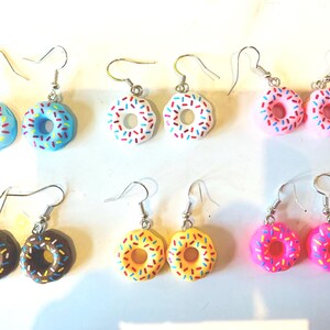 K&ouml;nnte beinhalten: Sechs Paar Ohrh&auml;nger, jeweils mit einem Mini-Donut-Design. Die Donuts sind in Blau, Wei&szlig;, Rosa, Braun, Gelb und Magenta gehalten, mit bunten Streuseln und silberfarbenen Ohrhaken.