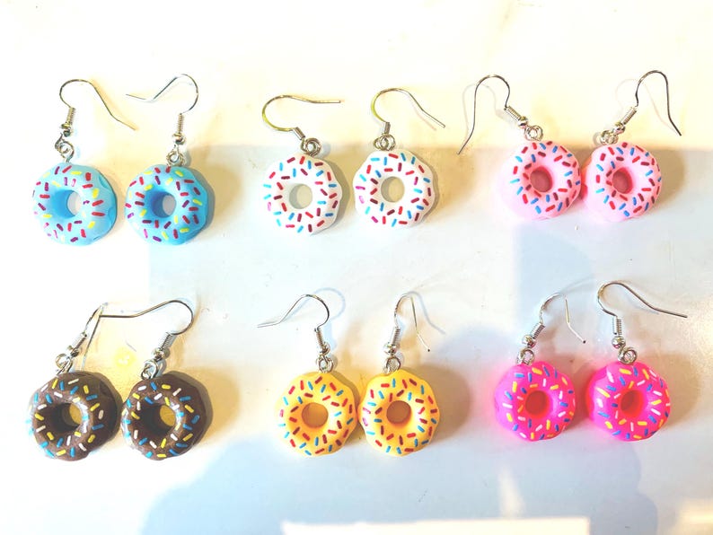 K&ouml;nnte beinhalten: Sechs Paar Ohrh&auml;nger, jeweils mit einem Mini-Donut-Design. Die Donuts sind in Blau, Wei&szlig;, Rosa, Braun, Gelb und Magenta gehalten, mit bunten Streuseln und silberfarbenen Ohrhaken.
