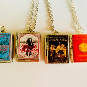 May include: Four miniature book pendant necklaces with silver chains. The books feature covers with titles like "Girl's Town", "Black Beauty", "Animal Farm", and "Outlander". Each book is a different color, including blue, red, and black.