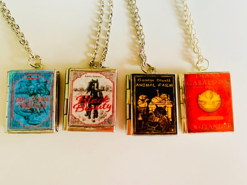 May include: Four miniature book pendant necklaces with silver chains. The books feature covers with titles like "Girl's Town", "Black Beauty", "Animal Farm", and "Outlander". Each book is a different color, including blue, red, and black.