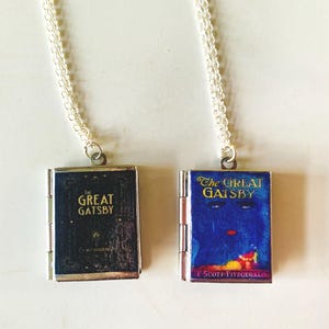May include: Two silver-toned necklaces each feature a miniature book pendant. One pendant is black with gold lettering that reads "The Great Gatsby." The other pendant is blue with the book title and a colorful illustration.