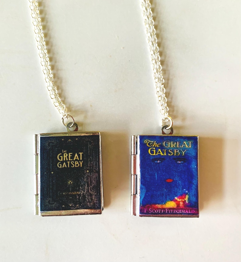 May include: Two silver-toned necklaces each feature a miniature book pendant. One pendant is black with gold lettering that reads "The Great Gatsby." The other pendant is blue with the book title and a colorful illustration.