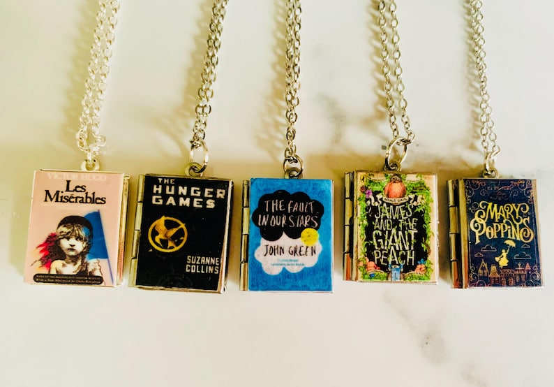 May include: Five silver necklaces each have a small book pendant. The pendants are miniature versions of book covers from popular novels: "Les Misérables", "The Hunger Games", "The Fault in Our Stars", "James and the Giant Peach", and "Mary Poppins".
