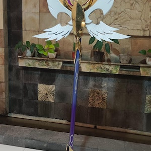 May include: A gold and purple staff with white wing-shaped accents and a star-shaped top. The staff has a detailed design with a gold base and a purple shaft. The top features a gold ring with a purple center.