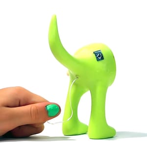 Pode incluir: Uma figurinha de cachorro de plástico verde com uma cauda longa. A cauda do cachorro está sendo puxada por uma mão com esmalte de unha verde.
