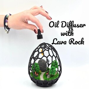 Peut inclure: Diffuseur d'huiles essentielles en fil de fer noir avec une roche volcanique et des arbres miniatures à l'intérieur. Le diffuseur a la forme d'un œuf et possède une petite ouverture en haut pour ajouter des huiles essentielles. Le texte "Diffuseur d'huiles essentielles avec roche volcanique" et "Pour les huiles essentielles" est écrit sous le diffuseur.