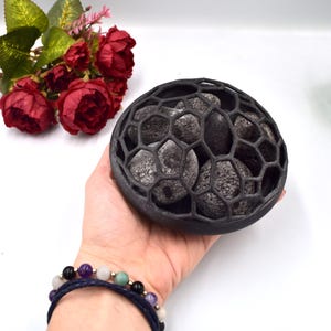 Op de afbeelding: Een zwarte keramische kom met een honingraatontwerp bevat een aantal zwarte lavstenen. De kom wordt in een hand met een blauwe armband vastgehouden.