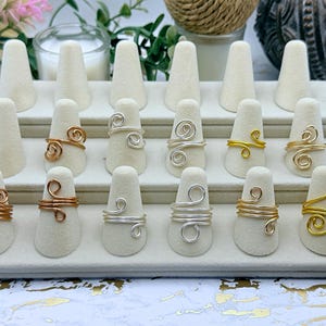 May include: A collection of twelve wire-wrapped rings displayed on a tiered beige velvet stand. The rings feature various metallic finishes including silver, gold, and copper tones, each designed with open-ended spiral or coiled wire patterns.