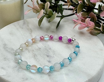Könnte beinhalten: Ein Armband mit weißen, irisierenden, rosa und blauen Perlen. Das Armband wird auf einer weißen Marmoroberfläche präsentiert, mit einer Kerze und Grünzeug im Hintergrund.