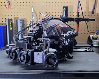 May include: A black tactical helmet featuring a multi-cam patterned fabric cover, a mounted flashlight on the side, and a front-facing array of three circular cooling fans. The helmet includes various adjustable straps, mounting hardware, and cables.