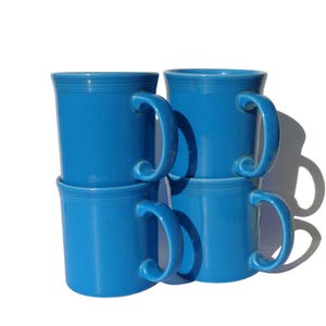 May include: A set of four bright blue ceramic mugs stacked in two columns. Each mug features a smooth, glossy finish, a cylindrical body with a slightly flared rim, and a curved handle attached to the side. The design includes subtle horizontal rings.