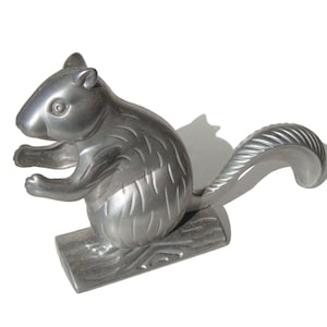 May include: A silver-colored squirrel nutcracker. The squirrel is in a sitting position, with its front paws raised. The tail curves upwards. The nutcracker is mounted on a wooden base.
