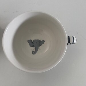 Puede incluir: Taza de t&eacute; de cer&aacute;mica blanca con una ilustraci&oacute;n de elefante gris en el centro. El asa presenta rayas azules y blancas. La taza est&aacute; vac&iacute;a y sobre una superficie blanca.