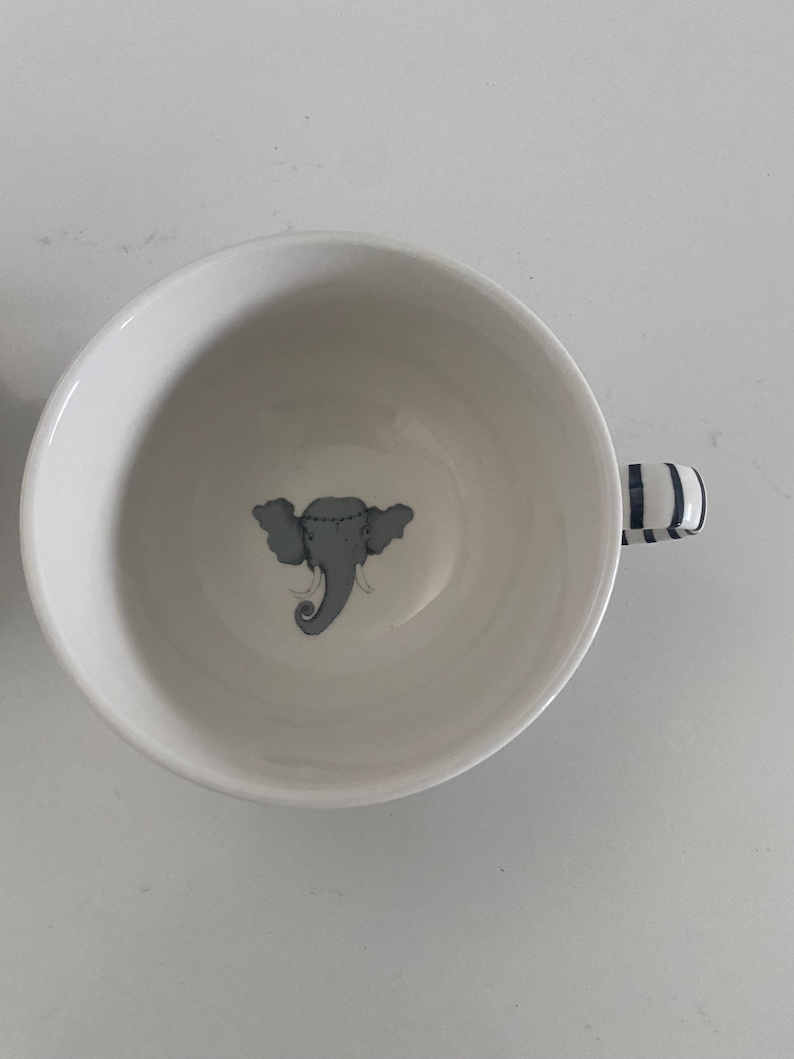 Puede incluir: Taza de t&eacute; de cer&aacute;mica blanca con una ilustraci&oacute;n de elefante gris en el centro. El asa presenta rayas azules y blancas. La taza est&aacute; vac&iacute;a y sobre una superficie blanca.