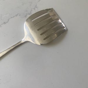 Peut inclure: Une spatule de cuisine en métal argenté dotée d'une tête large et plate avec quatre fentes rectangulaires horizontales. L'ustensile présente une surface lisse et réfléchissante ainsi qu'un manche fin partant de la base de la tête fendue.