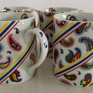 Puede incluir: Juego de cuatro tazas de cerámica blanca con un patrón de paisley en azul, amarillo, rojo y verde. Las tazas tienen un patrón de rayas diagonales con una raya amarilla, azul y roja.