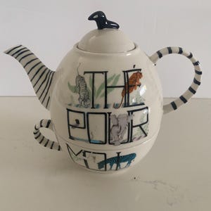 Puede incluir: Tetera de cer&aacute;mica color crema con asa y boquilla a rayas blancas y negras. La tetera presenta ilustraciones de tigres, cebras y un elefante, con las palabras "THE POOR MOM" impresas en la parte delantera. Una figura de p&aacute;jaro negro est&aacute; en la tapa.